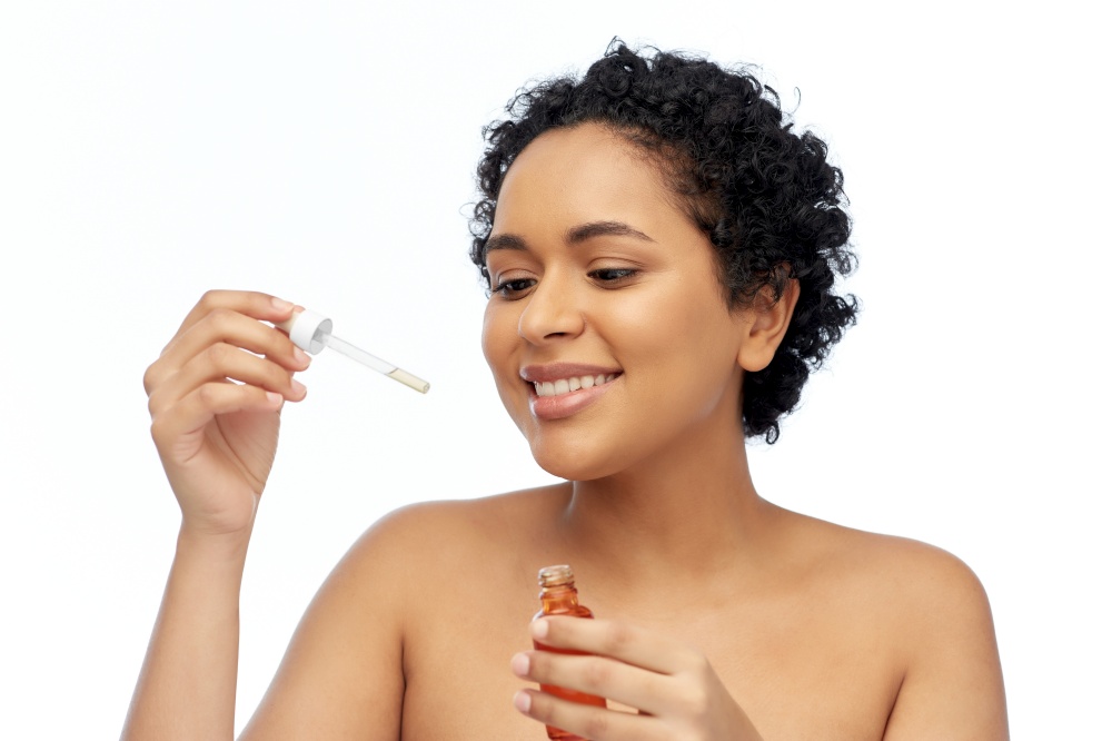beauty, cosmetics and skincare concept - portrait of happy smiling young african american woman with bare shoulders holding bottle of serum or face oil over white background. happy african american woman with bottle of serum