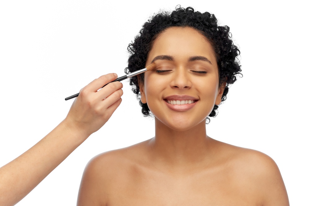 beauty, cosmetics and people concept - beautiful young african american woman and hand of make up artist with brush applying eye shadows over white background. happy woman and hand of make up artist with brush