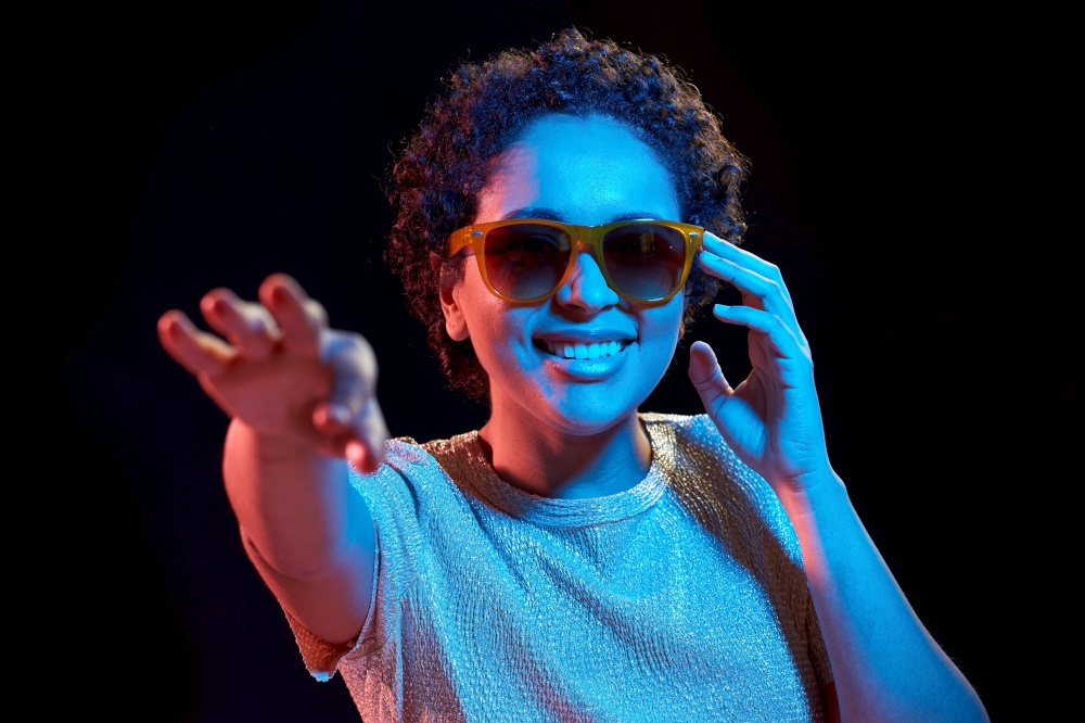 leisure, clubbing and nightlife concept - smiling young african american woman in sunglasses over ultraviolet neon lights on black background. african woman in sunglasses over neon lights