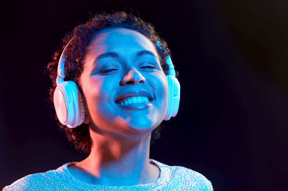 nightlife, technology and people concept - happy young african american woman in headphones listening to music and dancing in neon lights over black background. woman in headphones listening to music and dancing
