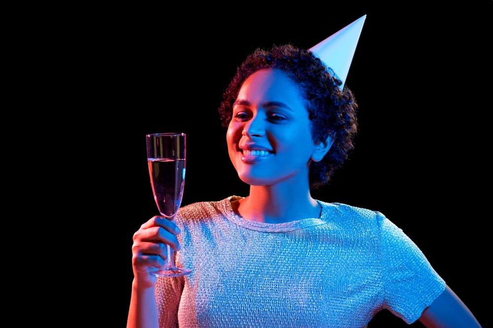 birthday, celebration and nightlife concept - happy smiling young african american woman in party cap with glass of champagne over ultraviolet neon lights on black background. african woman in party cap with glass of champagne