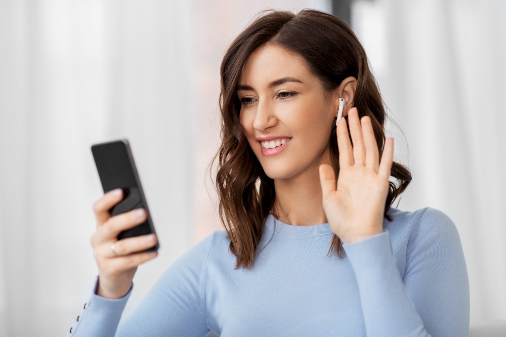 people, technology and leisure concept - happy young woman with wireless earphones and smartphone having video call and waving hand at home. woman with earphones and smartphone at home