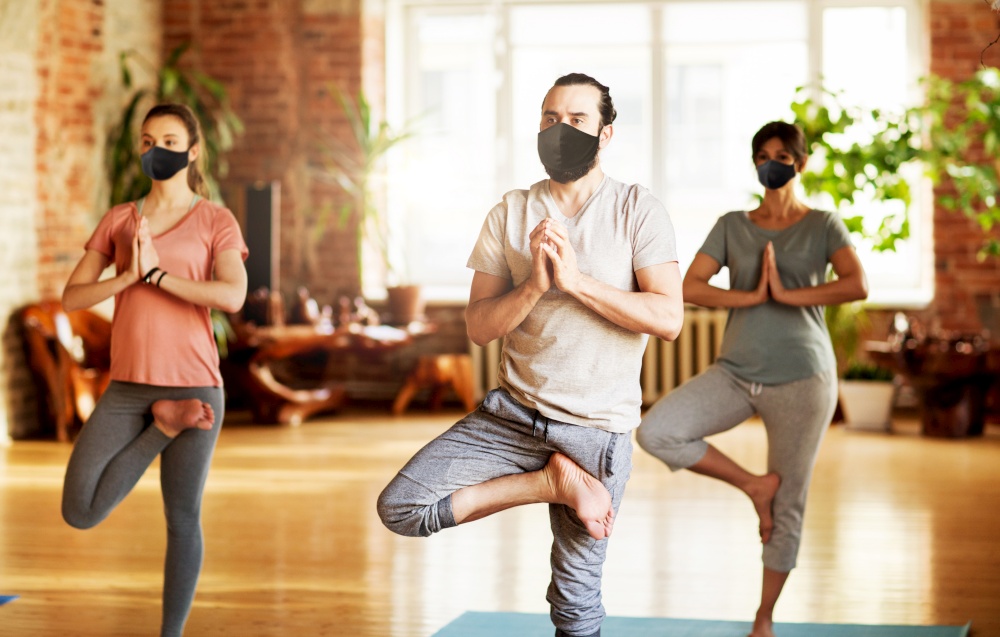 fitness, epidemic and health care concept - group of people wearing face protective black masks for protection from virus disease doing yoga in tree pose at studio. group of people in masks doing yoga at studio