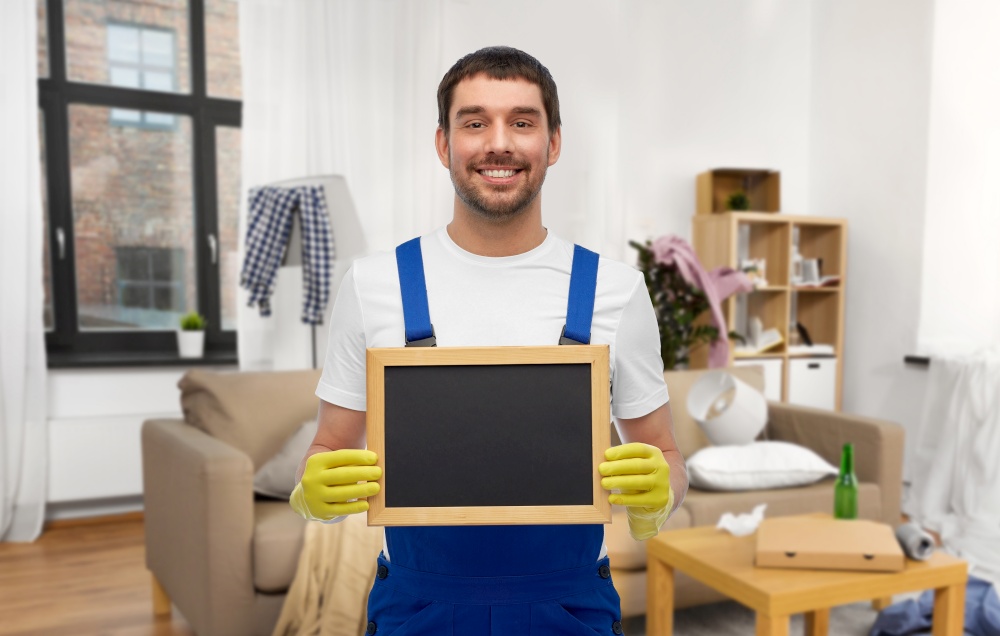 profession, cleaning service and people concept - happy smiling male worker or cleaner showing chalkboard over home room background. male worker or cleaner with chalkboard at home