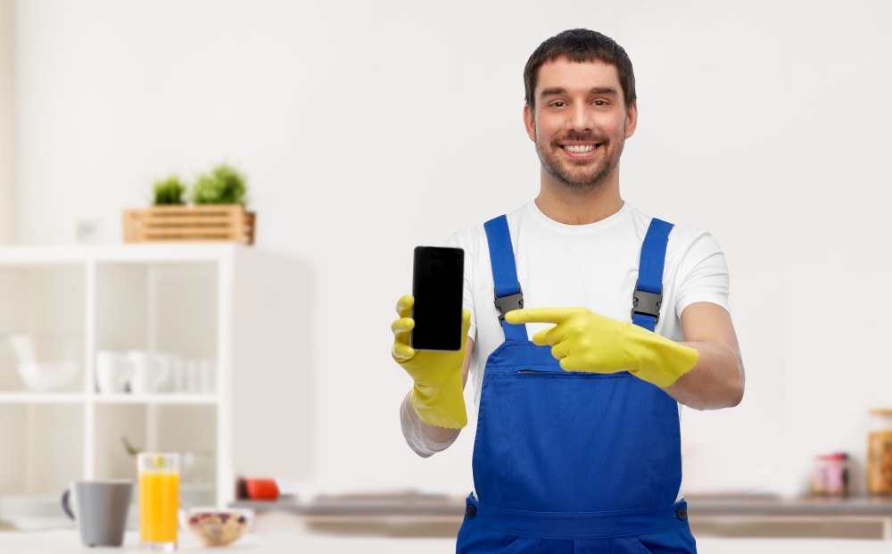 profession, cleaning service and plumbing concept - happy smiling male worker, plumber or cleaner in overall and gloves showing smartphone over home room background. male worker or cleaner with smartphone at kitchen