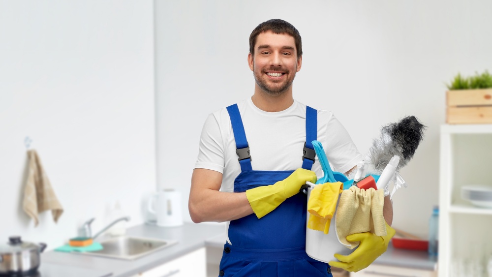 profession, service and people concept - happy smiling male worker or cleaner in overall and gloves with cleaning supplies over home room background. male cleaner with cleaning supplies at kitchen