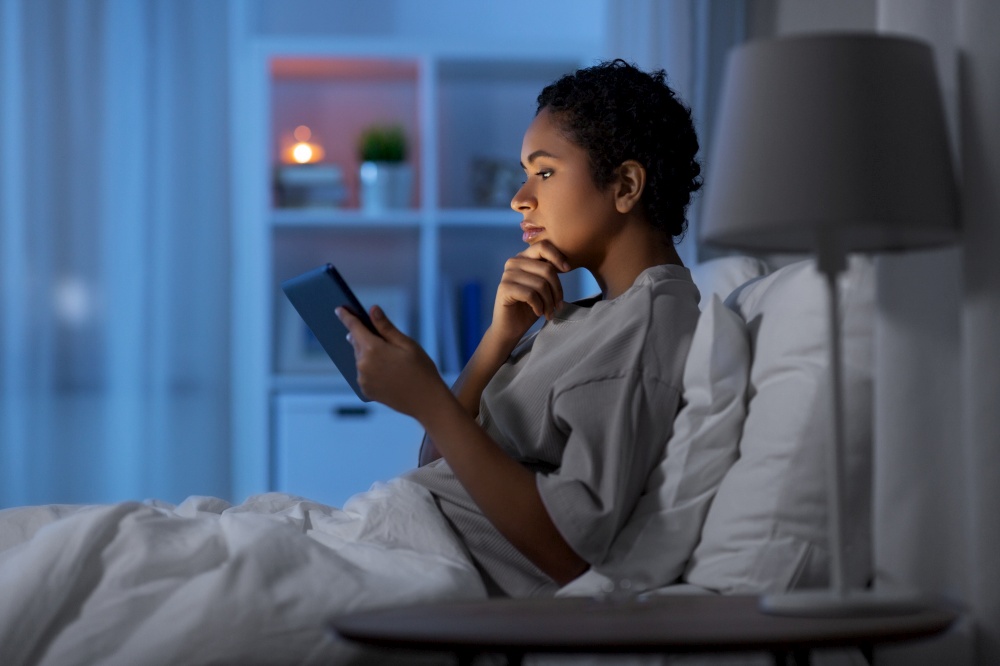 technology, internet and people concept - young african american woman with tablet pc computer lying in bed at home at night. woman with tablet pc in bed at home at night