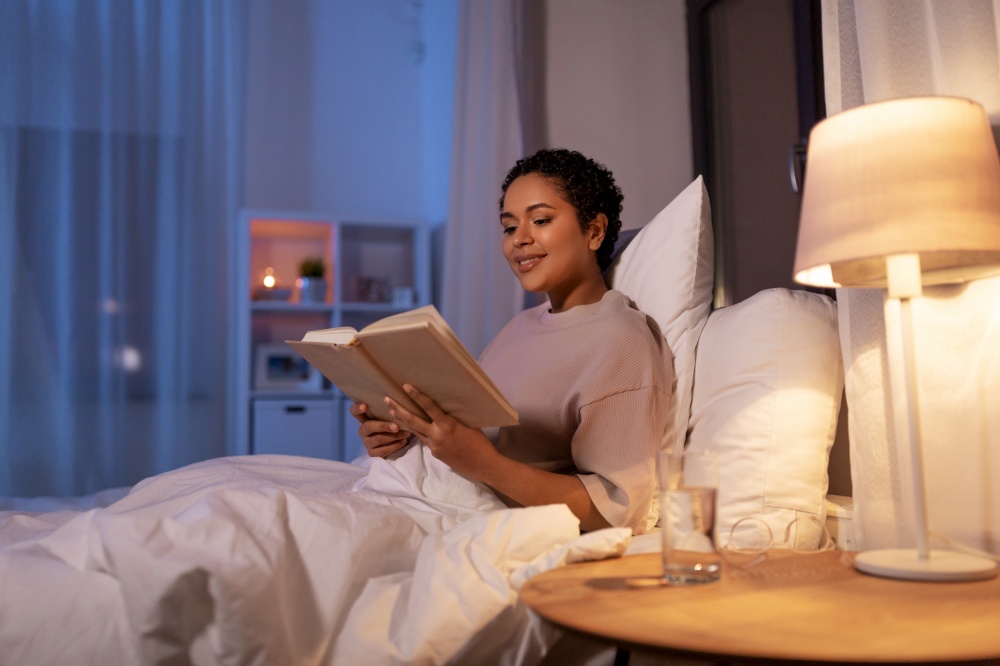 people, bedtime and rest concept - happy smiling young african american woman reading book in bed at home at night. smiling young woman reading book in bed at home