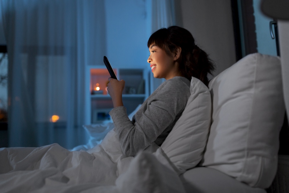technology, internet, communication and people concept - happy smiling young asian woman with smartphone lying in bed at home at night. asian woman with smartphone in bed at night