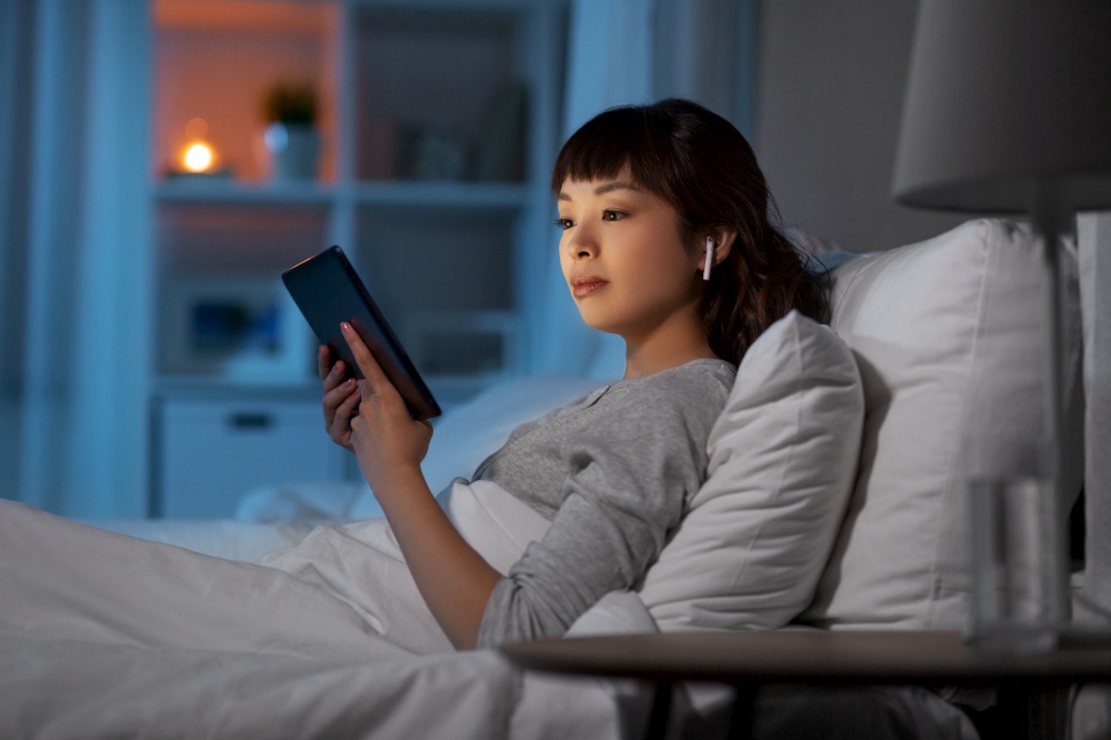 technology, internet and people concept - young asian woman with tablet pc computer and wireless earphones lying in bed at home at night. woman with tablet pc in earphones in bed at night