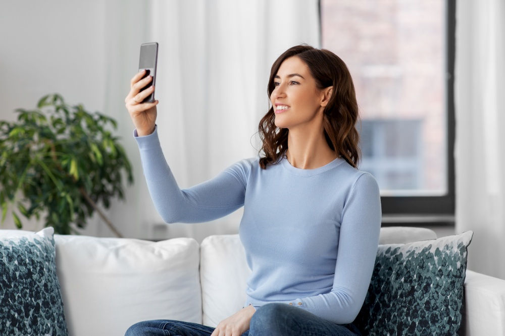 technology and people concept - happy smiling woman with smartphone and taking selfie sitting on sofa at home. happy woman with smartphone taking selfie at home
