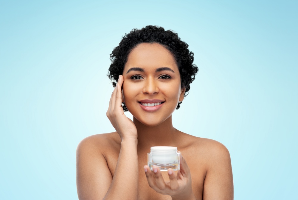 beauty and people concept - portrait of happy smiling young african american woman with bare shoulders holding jar of moisturizer over blue background. young african american woman with moisturizer