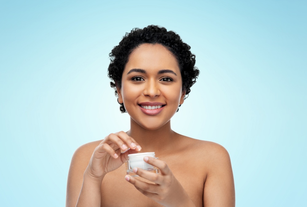beauty and people concept - portrait of happy smiling young african american woman with bare shoulders holding jar of moisturizer over blue background. young african american woman with moisturizer