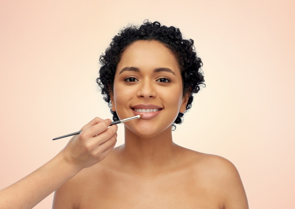 beauty, cosmetics and people concept - beautiful young african american woman and hand of make up artist with brush applying lipstick or lip gloss over beige background. happy woman and hand of make up artist with brush