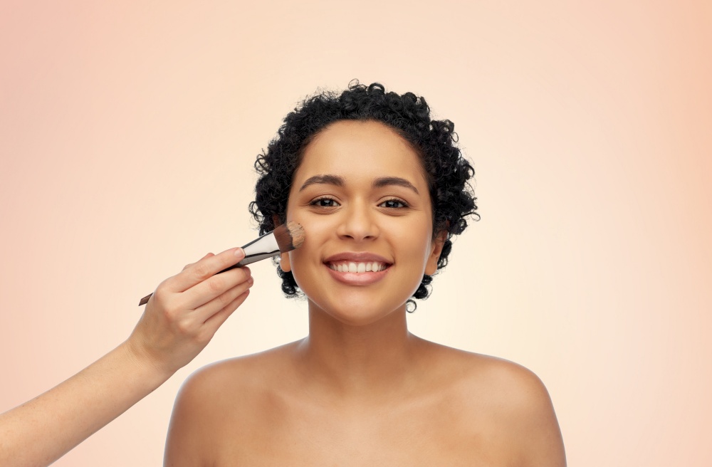 beauty, cosmetics and people concept - portrait of happy smiling young african american woman and hand of make up artist with brush applying foundation to her face over beige background. happy woman and hand of make up artist with brush