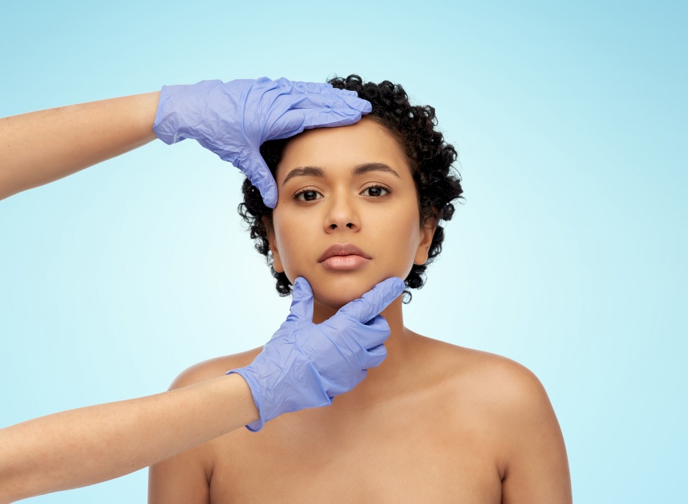beauty, plastic surgery and medicine concept - close up of face of beautiful young african american woman and hands of surgeon in gloves over blue background. face of african woman and hands in medical gloves