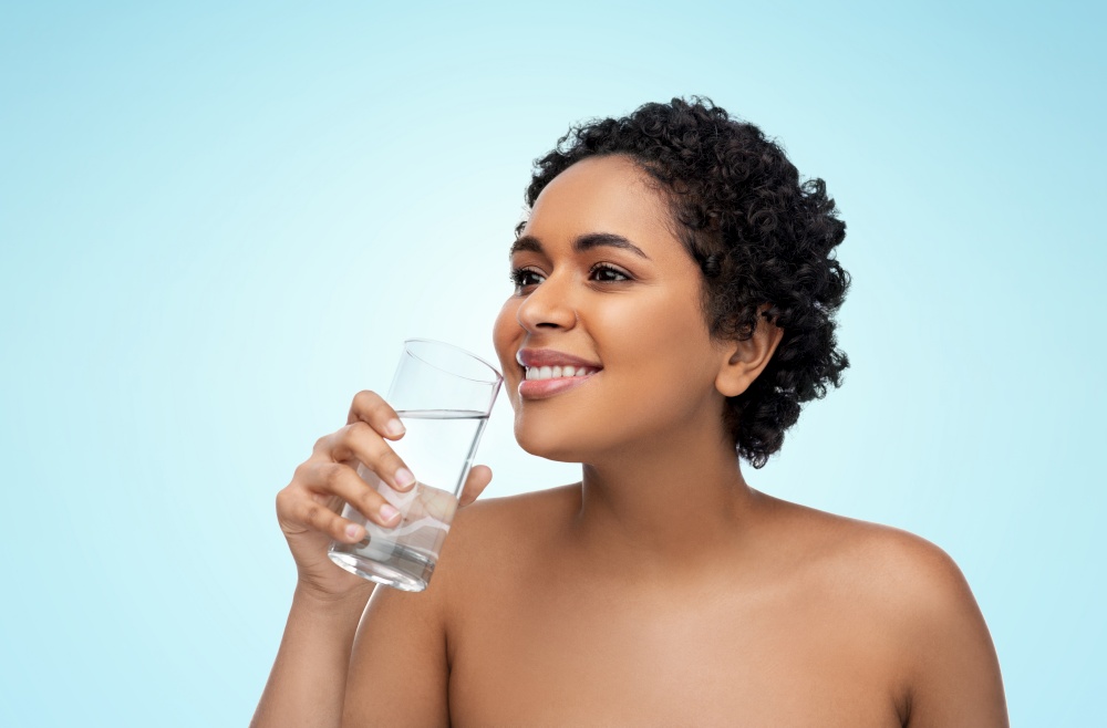 beauty and people concept - portrait of happy smiling young african american woman with bare shoulders with glass of water over blue background. young african american woman with glass of water
