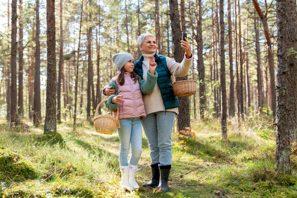 picking season, leisure and people concept - happy smiling grandmother and granddaughter with mushrooms in baskets taking selfie with smartphone in forest. grandma with granddaughter taking selfie in forest