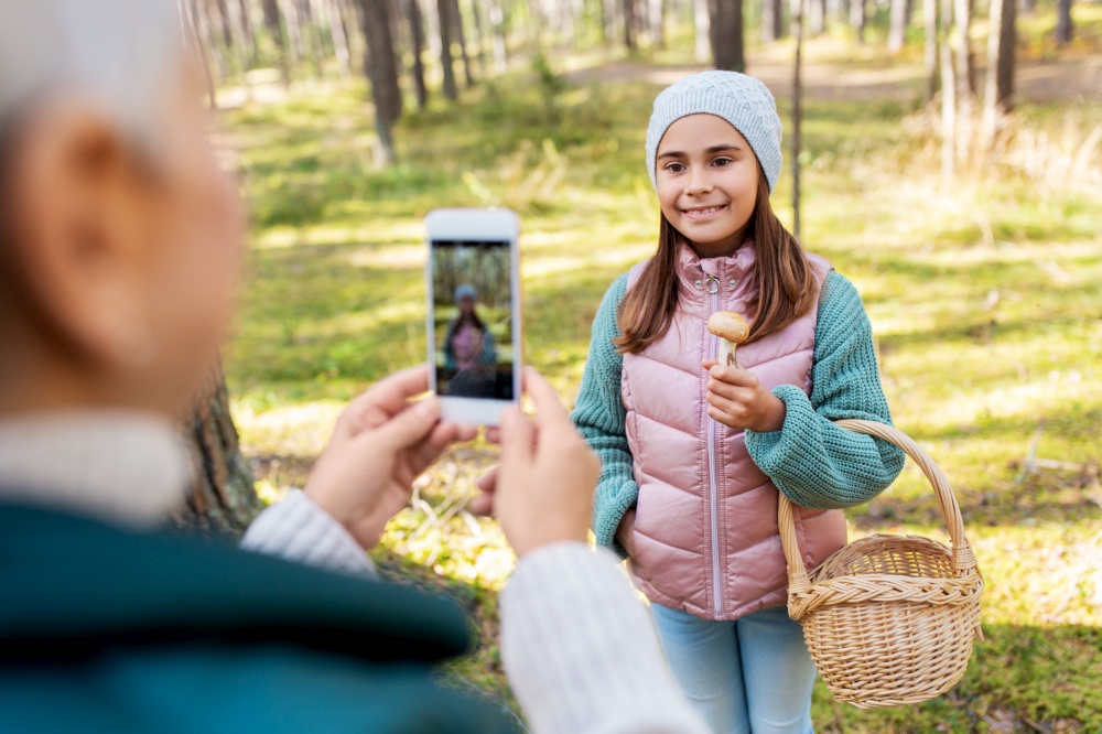 picking season, leisure and people concept - happy smiling grandmother with smartphone photographing granddaughter with mushrooms in basket in forest. grandma photographing granddaughter with mushrooms