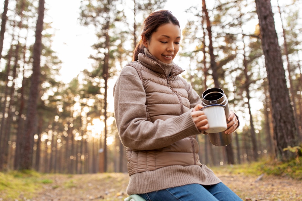 picking season, leisure and people concept - young asian woman with thermos and mug drinking tea in autumn forest. asian woman with thermos drinking tea in forest
