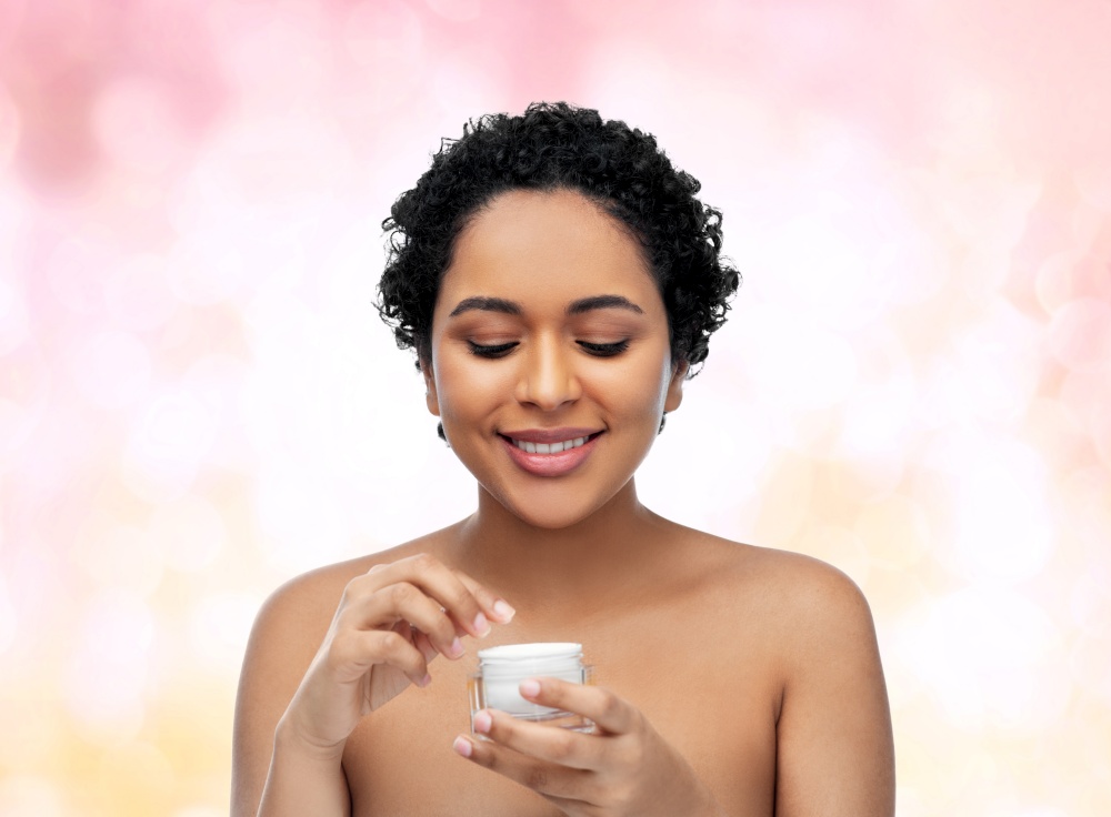 beauty, cosmetics and skincare concept - portrait of happy smiling young african american woman with bare shoulders holding jar of moisturizer over pink lights background. young african american woman with moisturizer