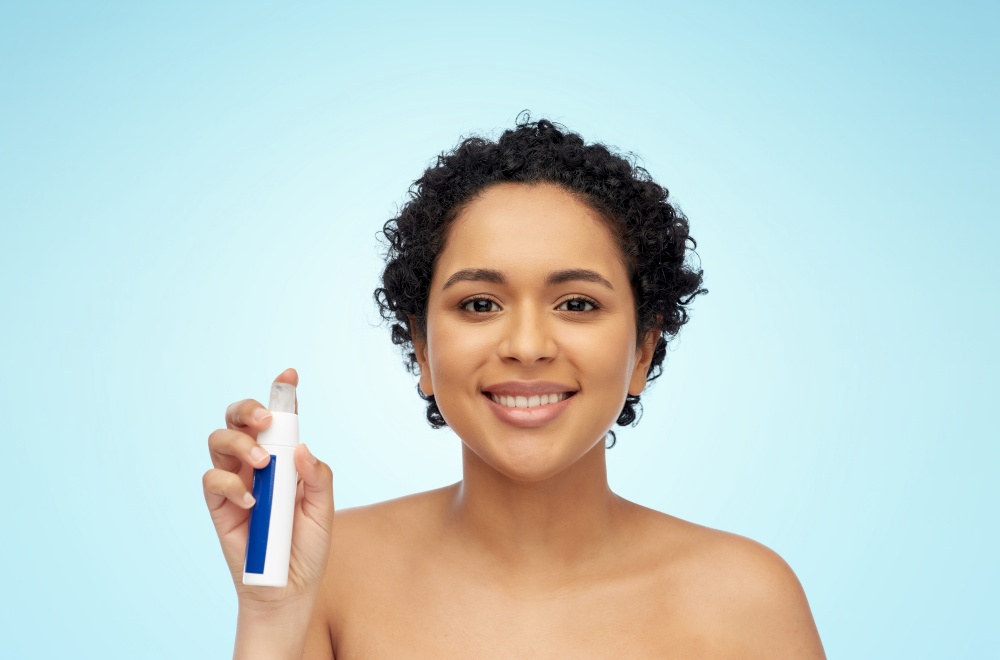 beauty, skin care and cosmetics concept - portrait of happy smiling young african american woman with bare shoulders with spray bottle over blue background. happy african woman with spray bottle