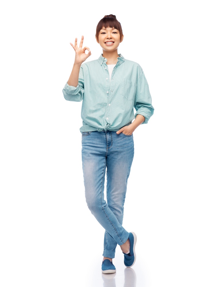people, ethnicity and gesture concept - happy asian young woman in cotton shirt and jeans showing ok hand sign over white background. happy asian woman over white background