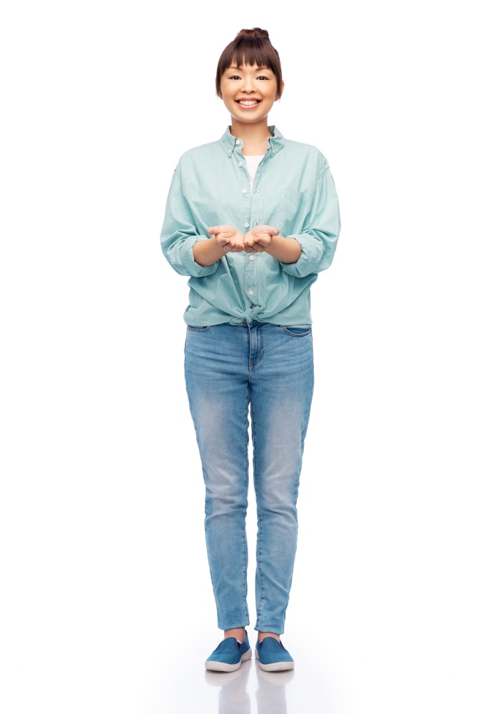 people, ethnicity and advertisement concept - happy asian young woman in cotton shirt and jeans holding something imaginary on her hands over white background. happy asian woman holding something on her hands