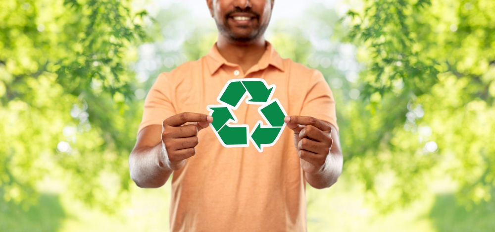 eco living, environment and sustainability concept - smiling young indian man in polo t-shirt holding green recycling sign over natural background. smiling indian man holding green recycling sign