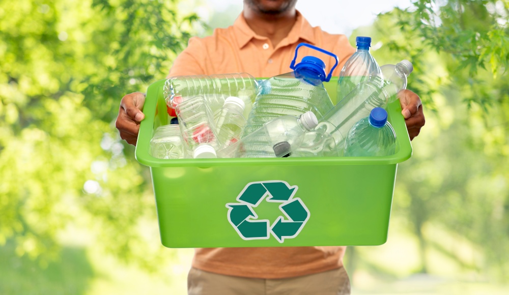 recycling, waste sorting and sustainability concept - close up of african american young man holding box with plastic bottles over green natural background. close up of young man sorting plastic waste