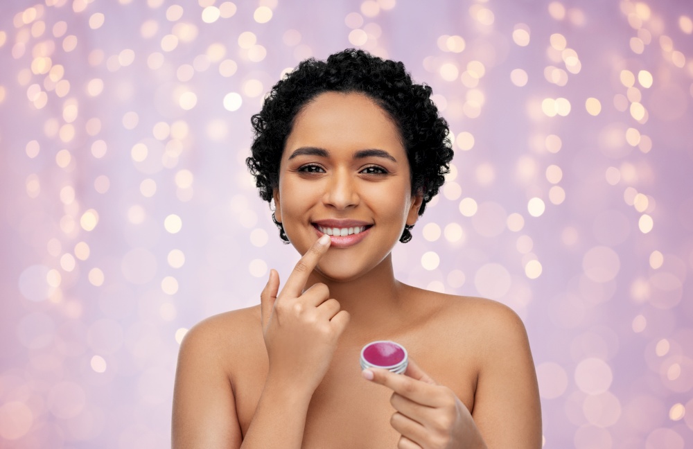 beauty, make up and cosmetics concept - beautiful young african american woman applying lip gloss with finger over festive lights on lilac background. african american woman applying lip gloss