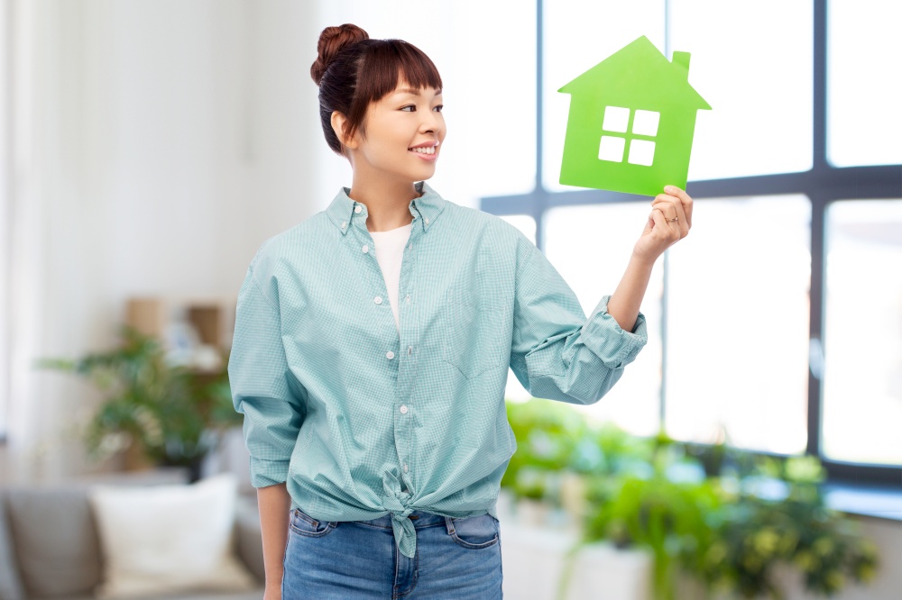 eco living, environment and sustainability concept - portrait of happy smiling young asian woman in turquoise shirt holding green house over home room background. smiling asian woman holding green house