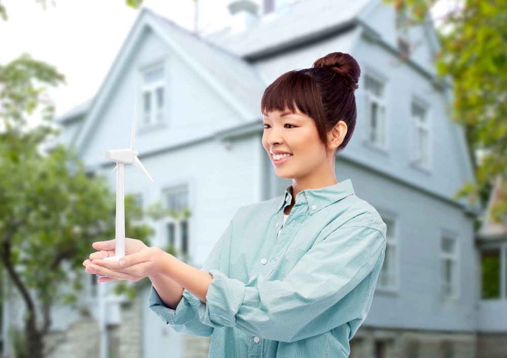 sustainable energy, power and people concept - happy smiling young asian woman with toy wind turbine over living house background. smiling young asian woman with toy wind turbine