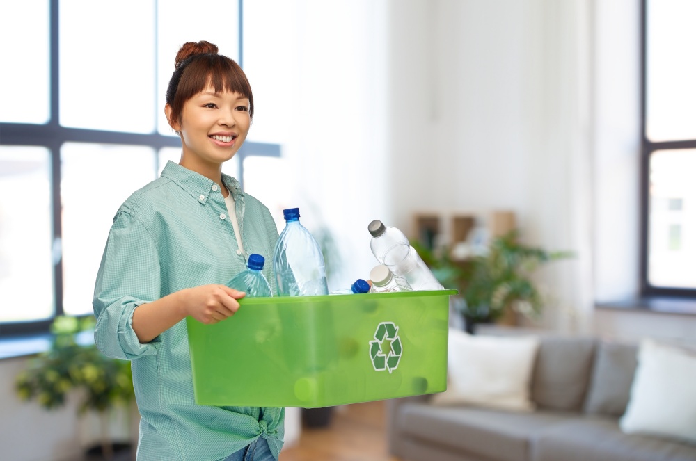recycling, waste sorting and sustainability concept - smiling young asian woman holding box with plastic bottles over home living room background. smiling young asian woman sorting plastic waste