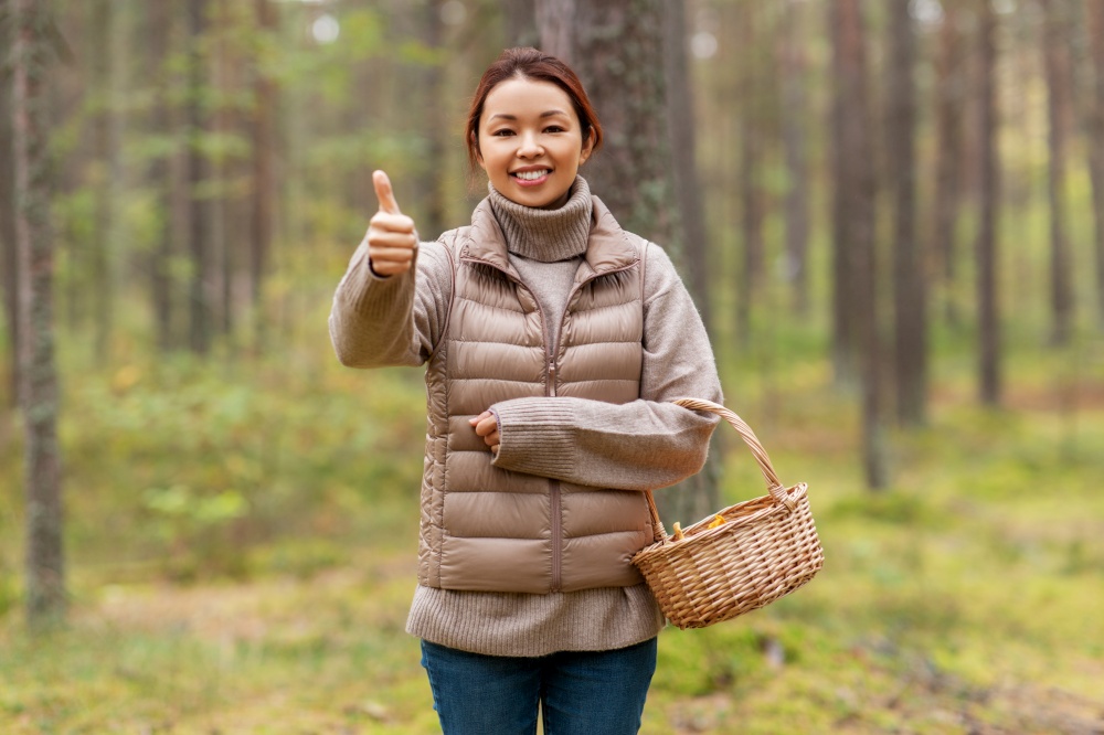 picking season, leisure and people concept - young asian woman with mushrooms in basket showing thumbs up in autumn forest. woman with mushrooms showing thumbs up in forest