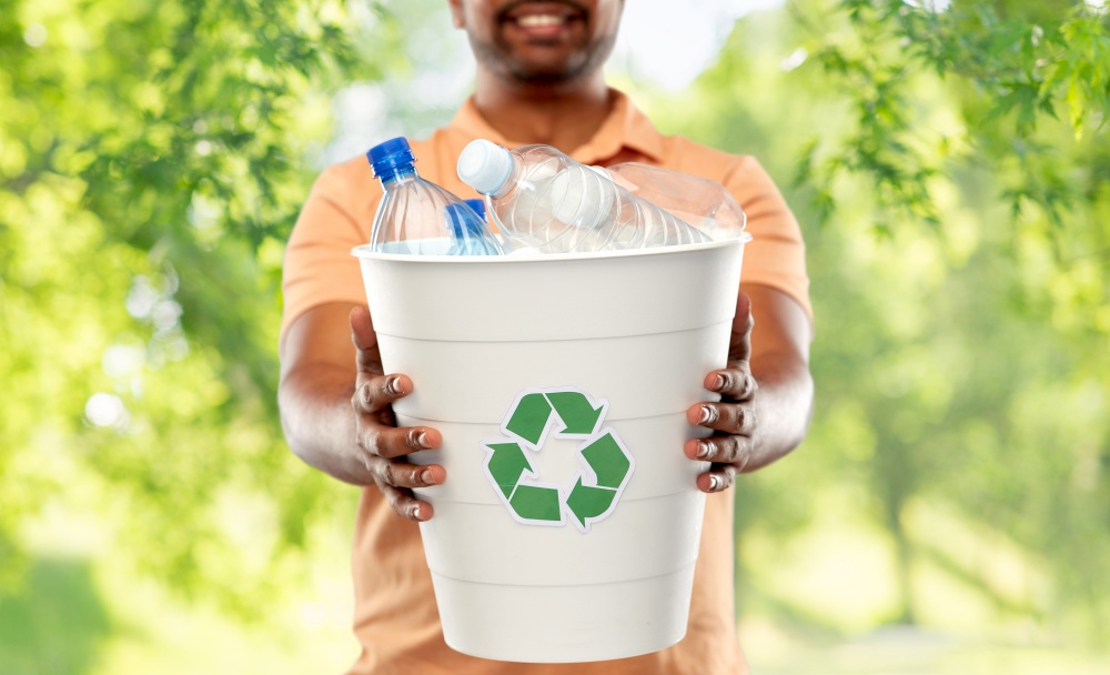 recycling, waste sorting and sustainability concept - close up of smiling young indian man holding bucket with plastic bottles over green natural background. close up of young indian man sorting plastic waste