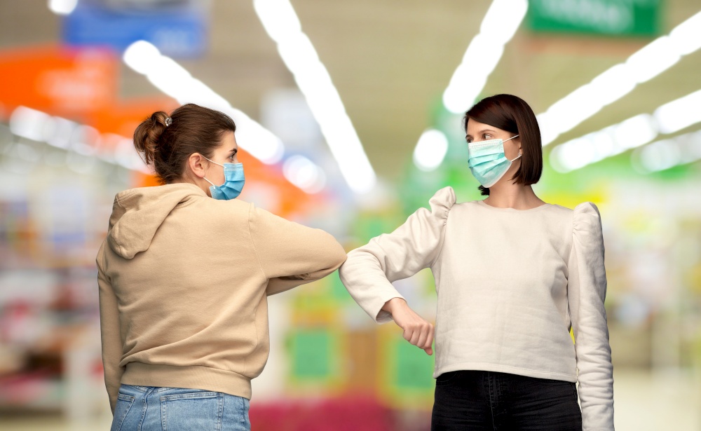 health protection, safety and pandemic concept - young women in masks meeting and making elbow bump greeting gesture over supermarket background. women in masks making elbow bump greeting gesture