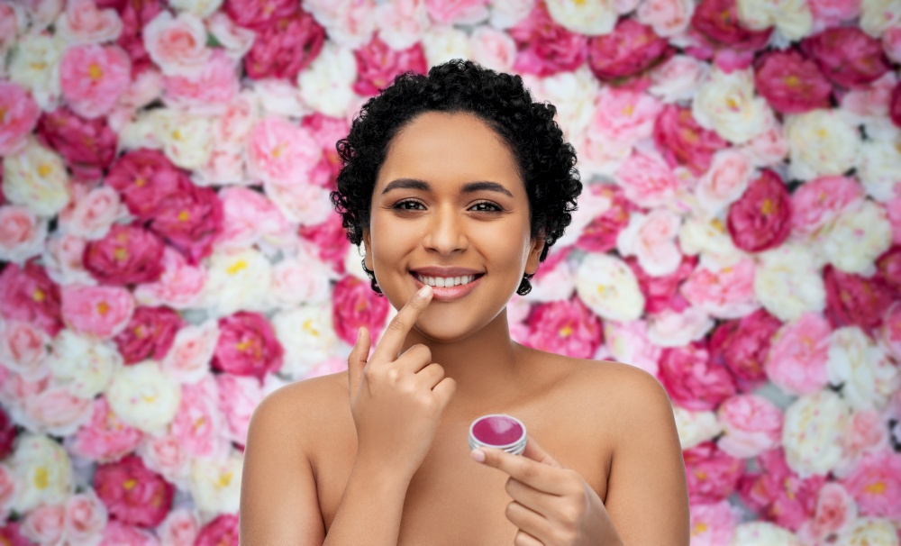 beauty, make up and cosmetics concept - beautiful young african american woman applying lip gloss with finger over flowers on background. african american woman applying lip gloss