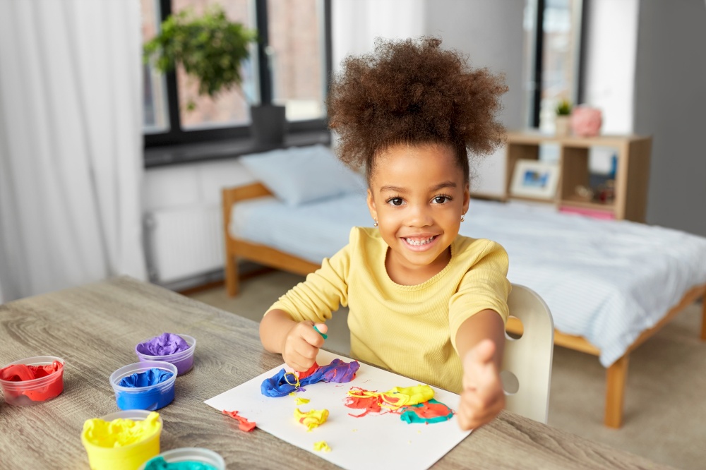 childhood, creativity and hobby concept - little african american girl playing with modeling clay and showing thumbs up at home. little girl with modeling clay playing at home
