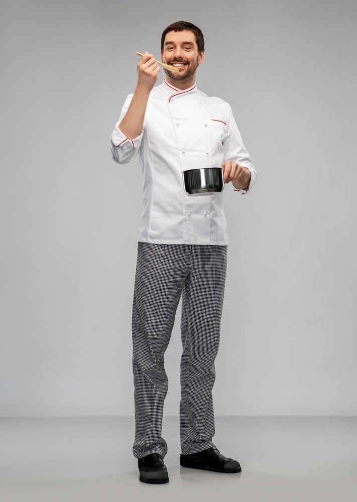 cooking, culinary and people concept - happy smiling male chef in jacket with pot or saucepan and spoon tasting food over grey background. happy smiling male chef with saucepan tasting food