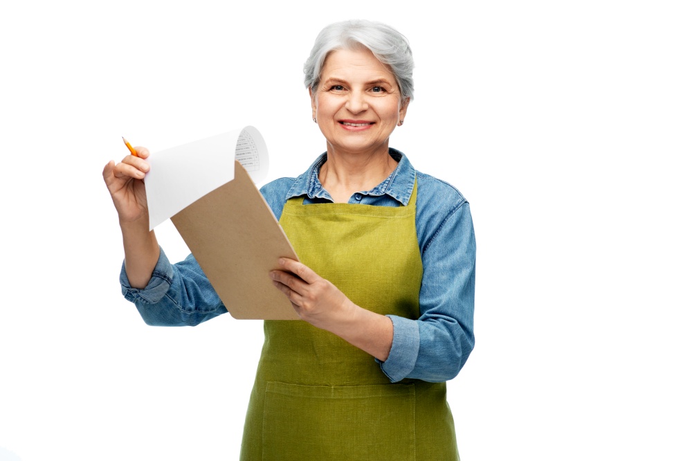 gardening, farming and old people concept - portrait of smiling senior woman in green garden apron with clipboard over white background. smiling old woman in garden apron with clipboard