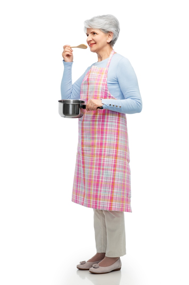 food cooking, culinary and old people concept - portrait of smiling senior woman in kitchen apron with pot and spoon over white background. senior woman in apron with pot cooking food