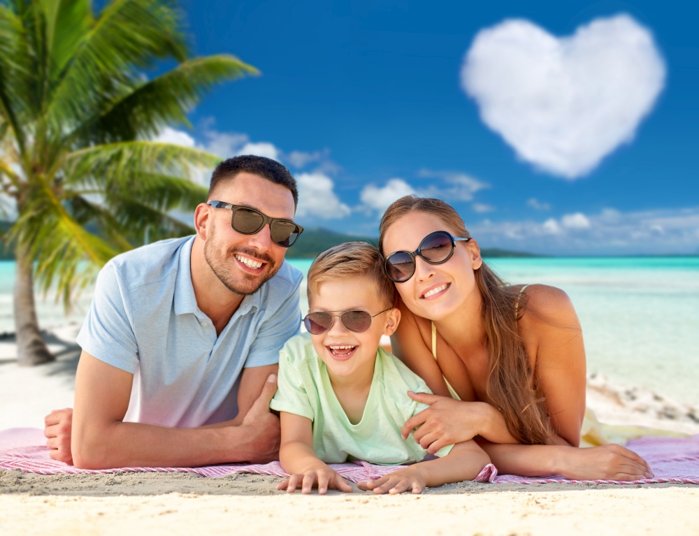 family, travel and tourism concept - happy mother, father and little son in sunglasses lying on blanket over tropical beach in french polynesia and heart shaped cloud on background. happy family lying over tropical beach background
