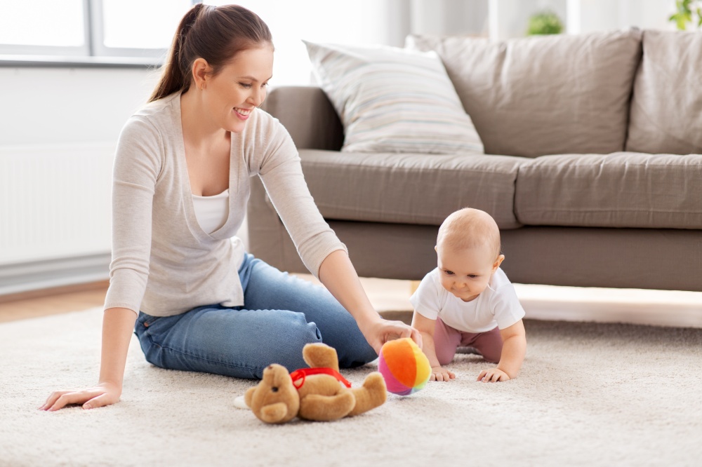 family, motherhood and people concept - happy smiling mother and little baby daughter playing with soft ball toy and teddy bear on floor at home. happy mother playing with little baby at home