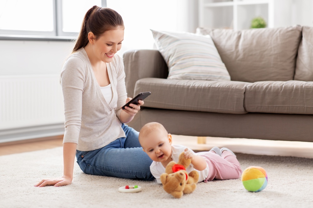 family, motherhood and people concept - happy smiling mother with smartphone and little baby daughter playing with soft ball toy and teddy bear on floor at home. mother with smartphone and baby playing at home
