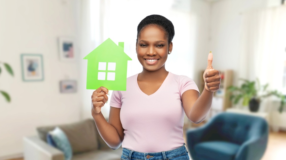 eco living, environment and sustainability concept - happy smiling young african american woman holding green house showing thumbs up over home room background. smiling african american woman holding green house