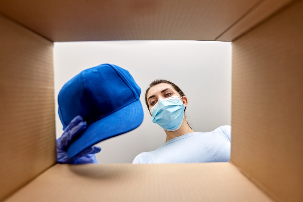 home delivery, shipping and pandemic concept - woman in protective medical mask and gloves opening parcel box, looking inside and taking clothes out. woman in mask opening parcel box with clothes