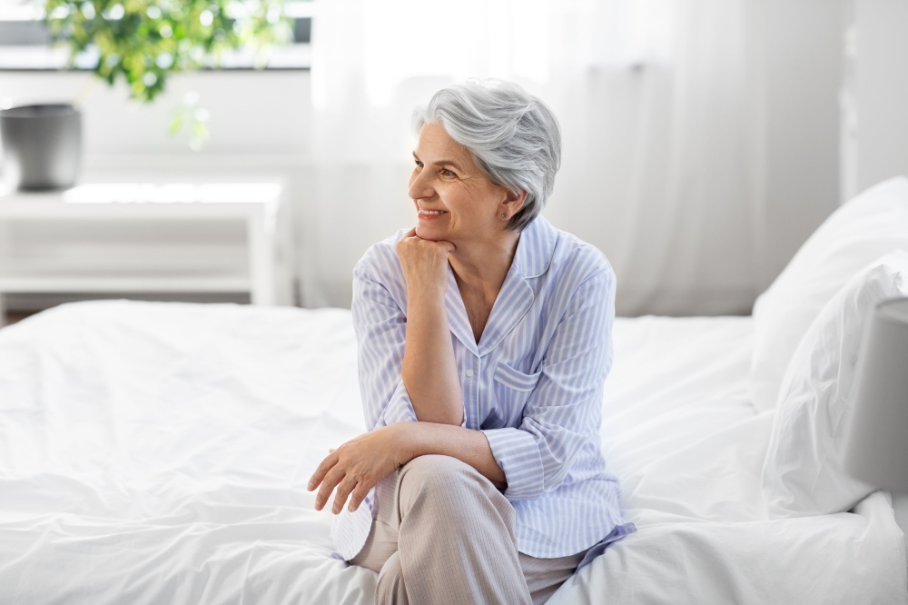 old age and people concept - happy smiling senior woman in pajamas sitting on bed at home bedroom. happy senior woman sitting on bed at home bedroom