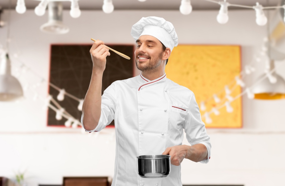 cooking, culinary and people concept - happy smiling male chef in toque with pot or saucepan and spoon tasting food over restaurant background. happy smiling male chef with saucepan tasting food
