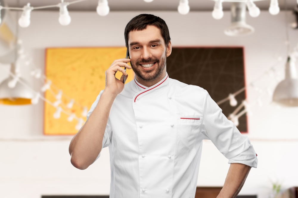 cooking, culinary and people concept - happy smiling male chef in jacket calling on smartphone over restaurant background. happy smiling male chef calling on smartphone